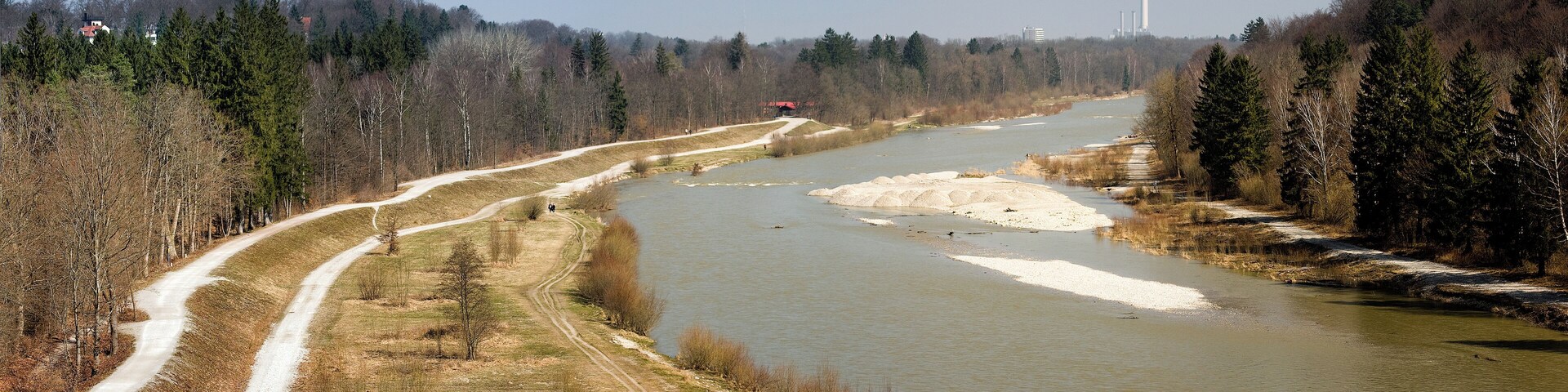 A river with a dirt path alongside it, surrounded by trees and a distant industrial facility.
