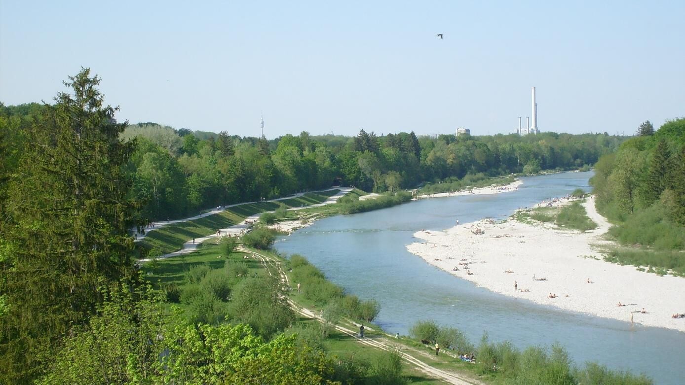 A river winding through a forested area with a sandy beach and industrial structures in the background.