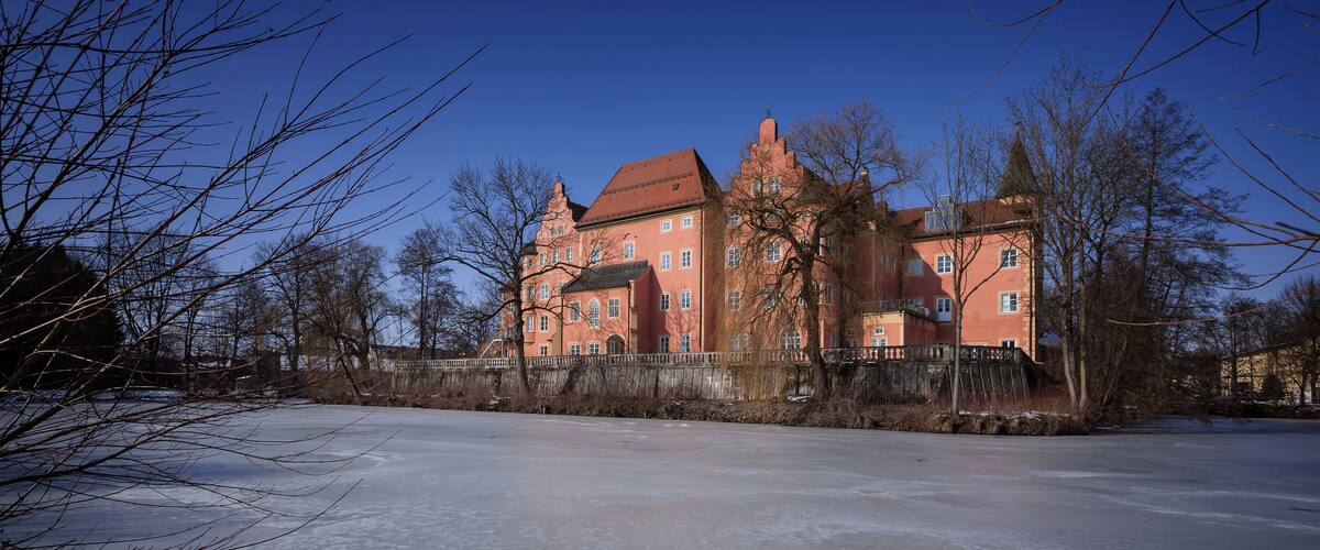 A large, historic red building with multiple towers and a surrounding frozen lake.