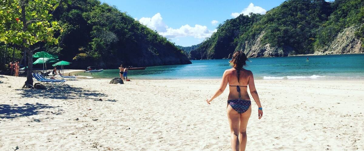 A woman walking on a sandy beach with a clear blue sky and lush green hills in the background.