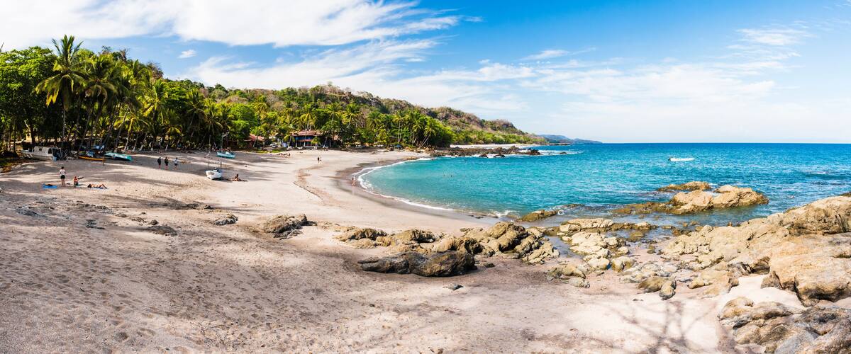 A tropical beach with clear turquoise waters, rocky shoreline, and palm trees.