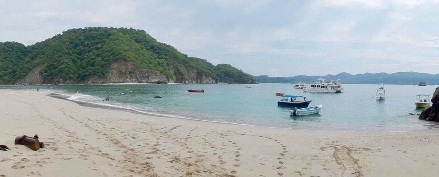 A beach with footprints, boats, and a forested hill in the background.
