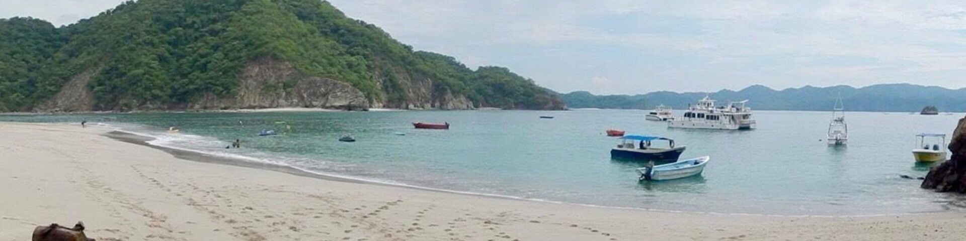 Uma praia com pegadas, barcos e uma colina arborizada ao fundo.
