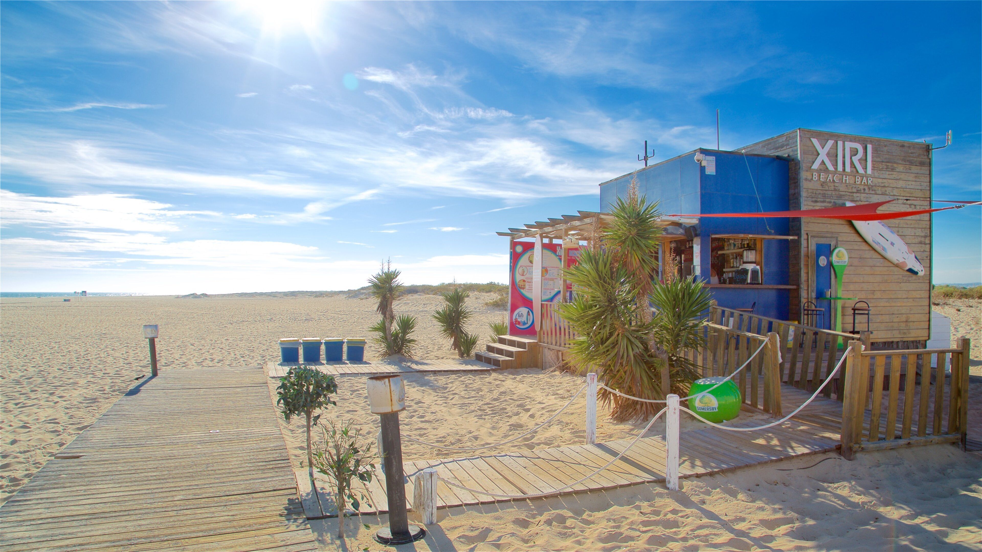 Un bar de playa llamado XIRI Beach Bar con estructura de madera, palmeras y un paseo de madera que lleva a la playa.