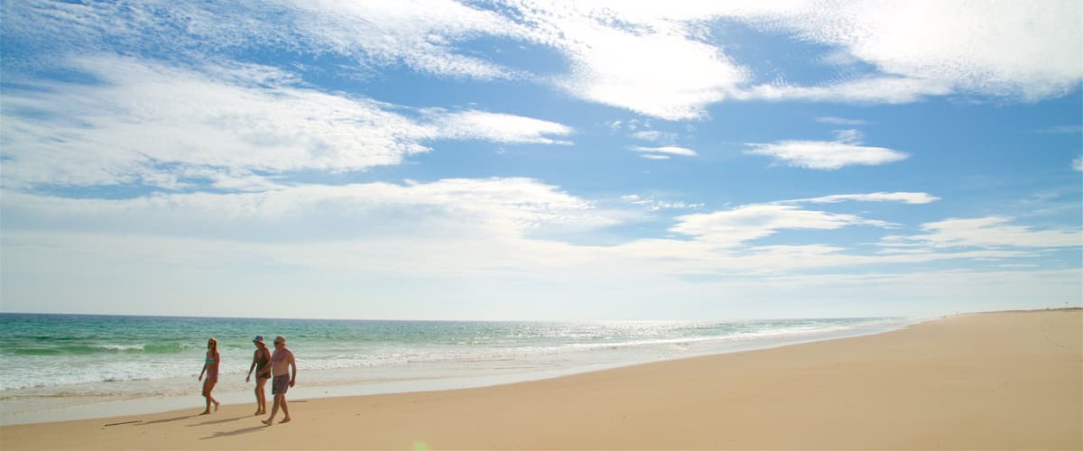 A beach with people walking along the shore, a clear sky with scattered clouds, and the ocean in the background.