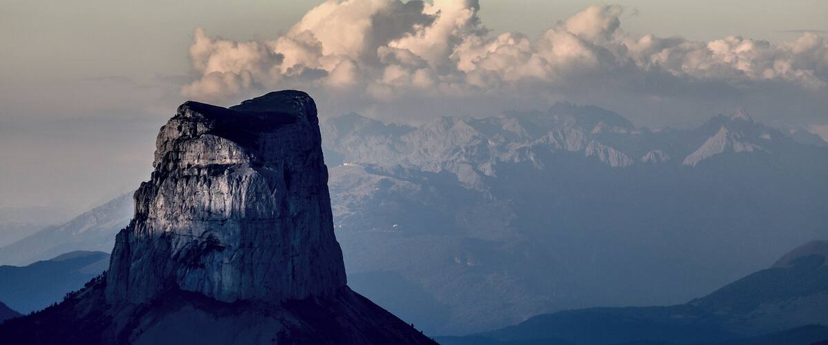 A mountain peak with a prominent rocky formation in the foreground and a range of snow-capped mountains in the background.