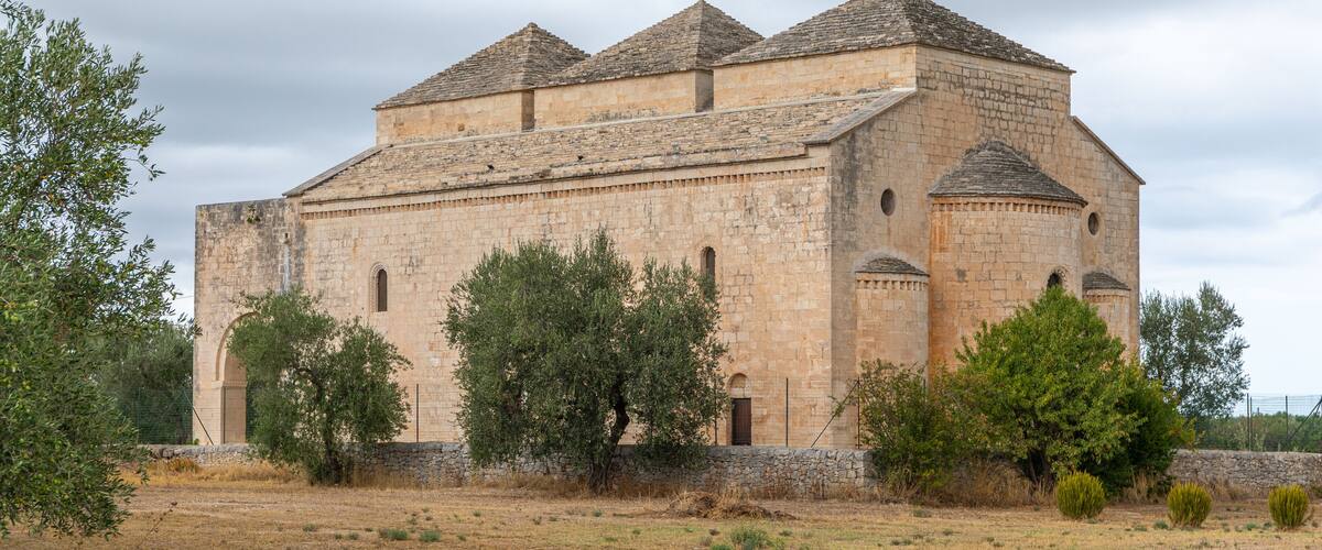 A historic stone building with a tiled roof, surrounded by olive trees and dry grass.