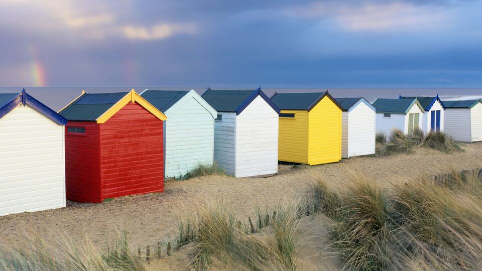 A row of colorful beach huts along a sandy beach with dunes and a stormy sky.