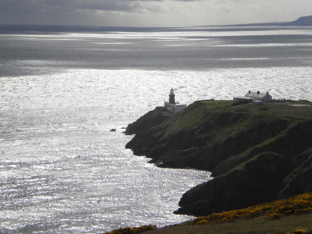 A lighthouse on a rocky coastline with a view of the sea and distant hills.