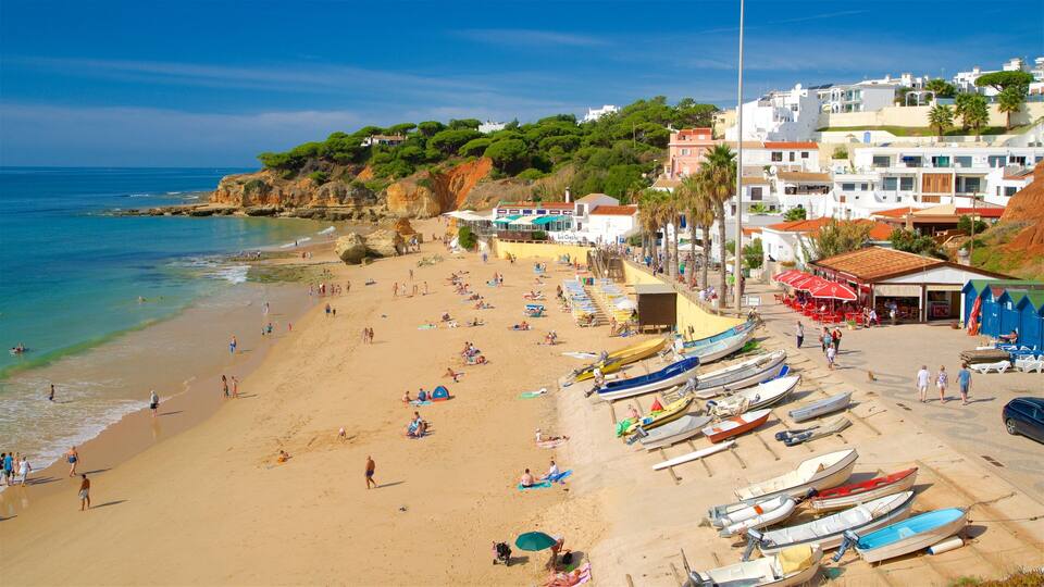A beach with people sunbathing and swimming, boats lined up on the shore, and buildings in the background.