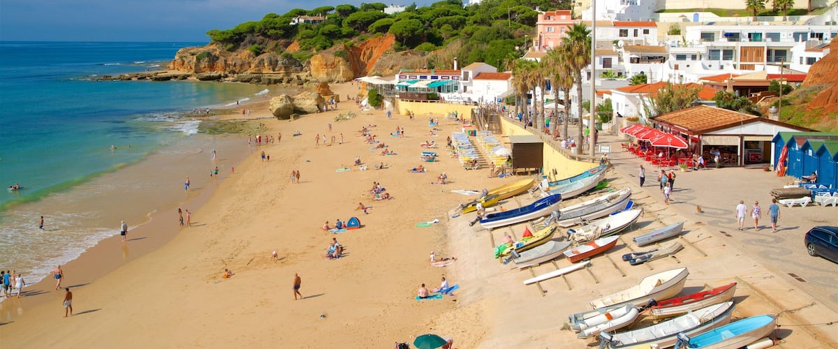 A beach with people sunbathing and swimming, boats lined up on the shore, and buildings in the background.