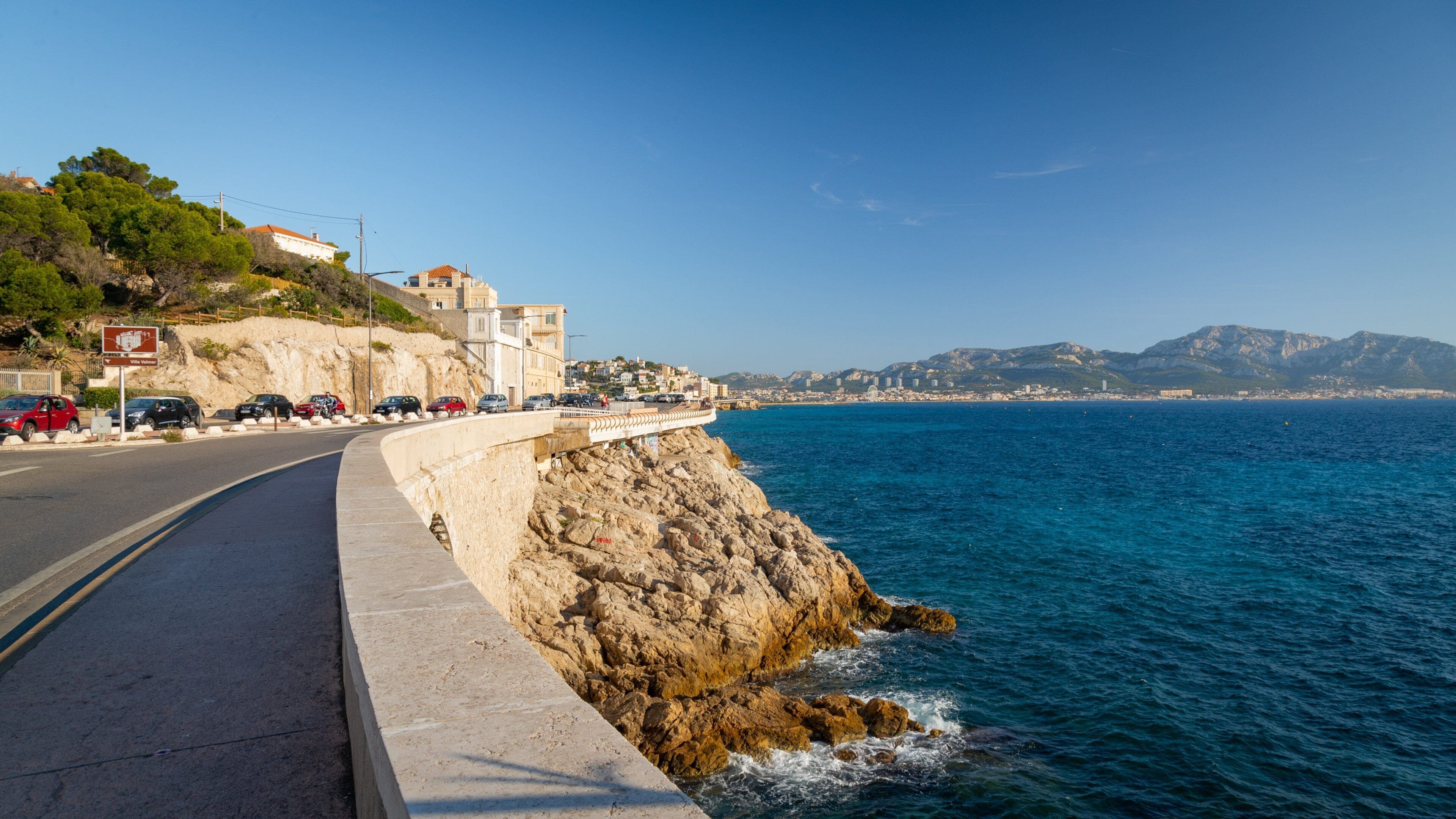 A coastal road with a stone barrier, parked cars, and a cityscape in the background.