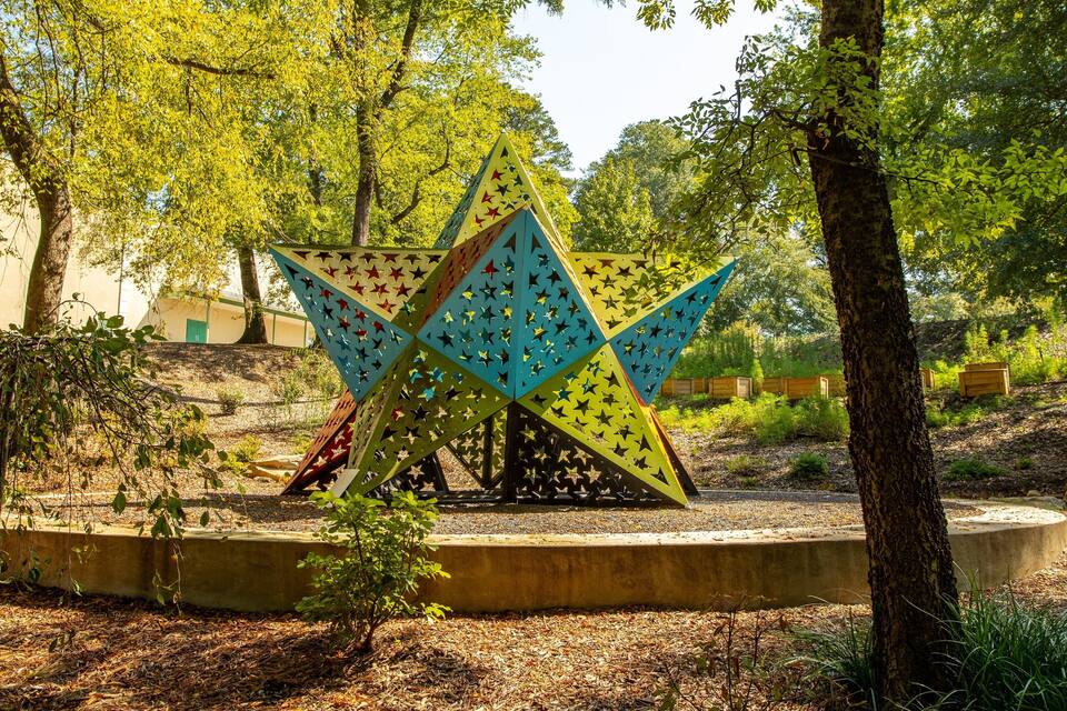 A colorful star-shaped sculpture in a park with trees and wooden structures in the background.