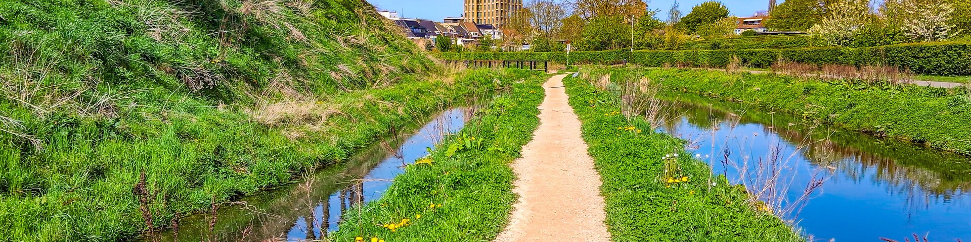 A canal with a dirt path, green vegetation, and a tall building in the background.