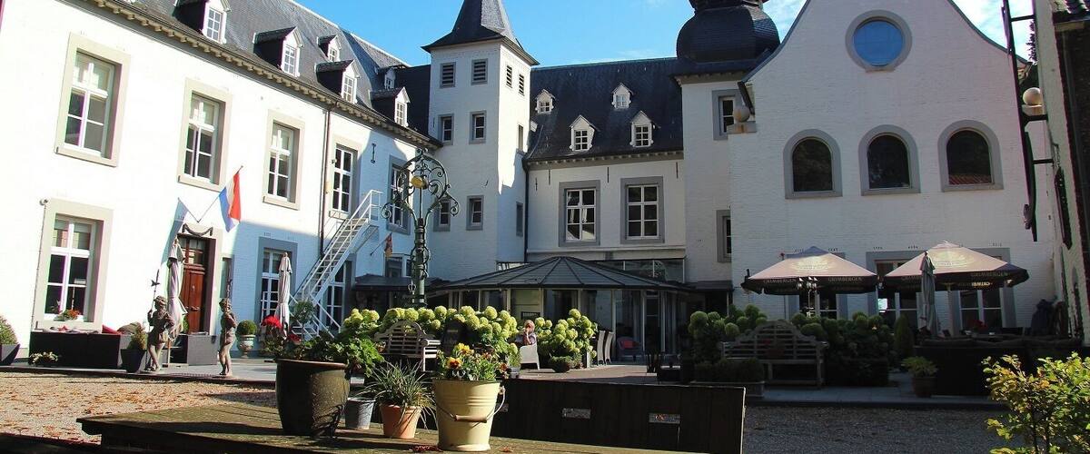 A courtyard with a white building featuring a tall spire, a smaller building with a round window, and a bench area with potted plants.
