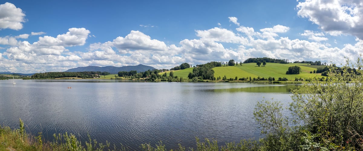 A calm lake surrounded by green hills and a clear blue sky with scattered clouds.