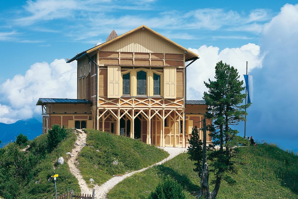 A wooden house with a steep roof and a balcony, surrounded by a mountainous landscape and a clear sky.