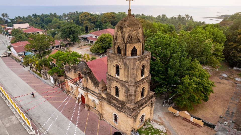 A church with a bell tower and a cross on top, surrounded by trees and a coastal view.