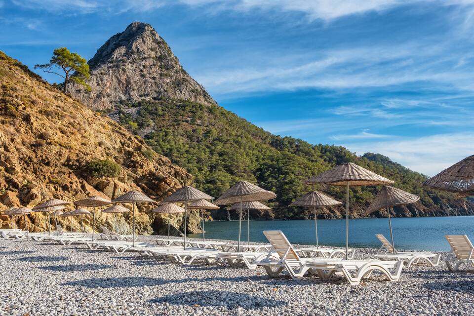 A pebble beach with lounge chairs and thatched umbrellas, a rocky mountain, and a calm sea.