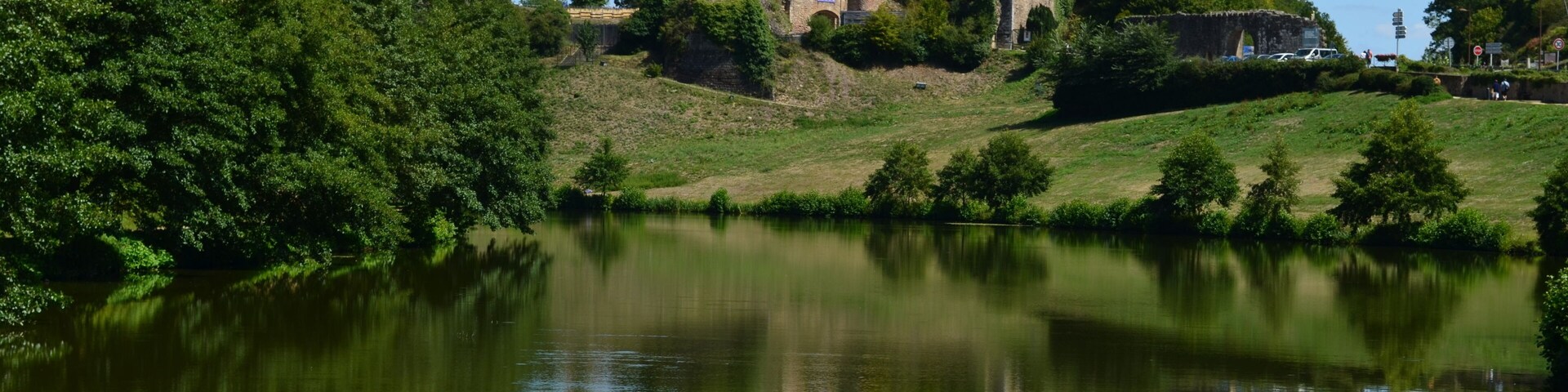 A castle on a hill overlooking a calm river, surrounded by lush greenery.