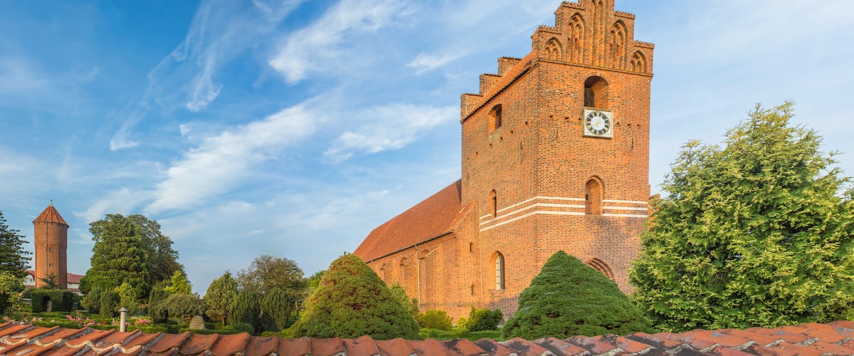 A historic brick church with a clock tower, surrounded by lush greenery and a clear blue sky.