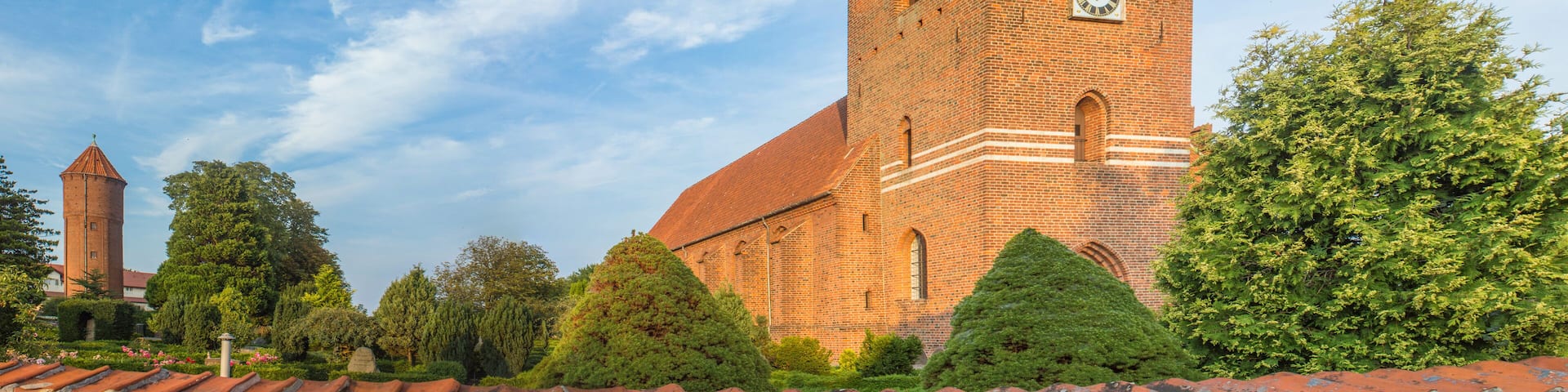 A historic brick church with a clock tower, surrounded by lush greenery and a clear blue sky.