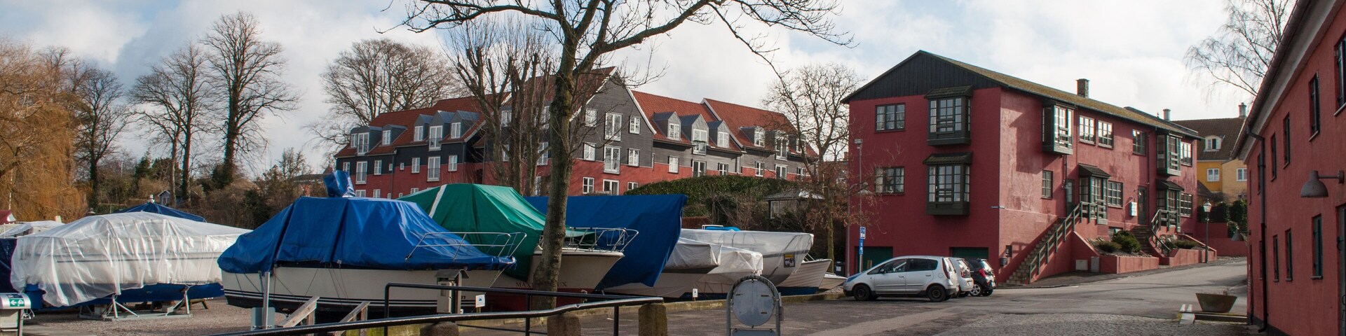 Ruas de paraleipípedos com barcos cobertos por lonas azuis, um edifício vermelho e uma árvore sem folhas.
