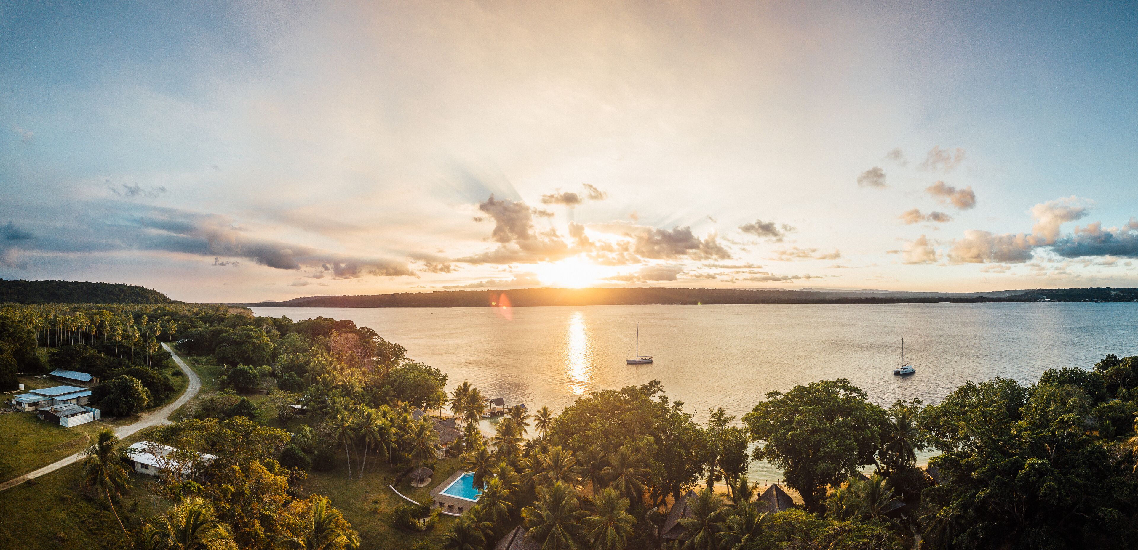 Um pôr do sol sobre um lago tranquilo, com barcos, cercado por vegetação exuberante e alguns edifícios.
