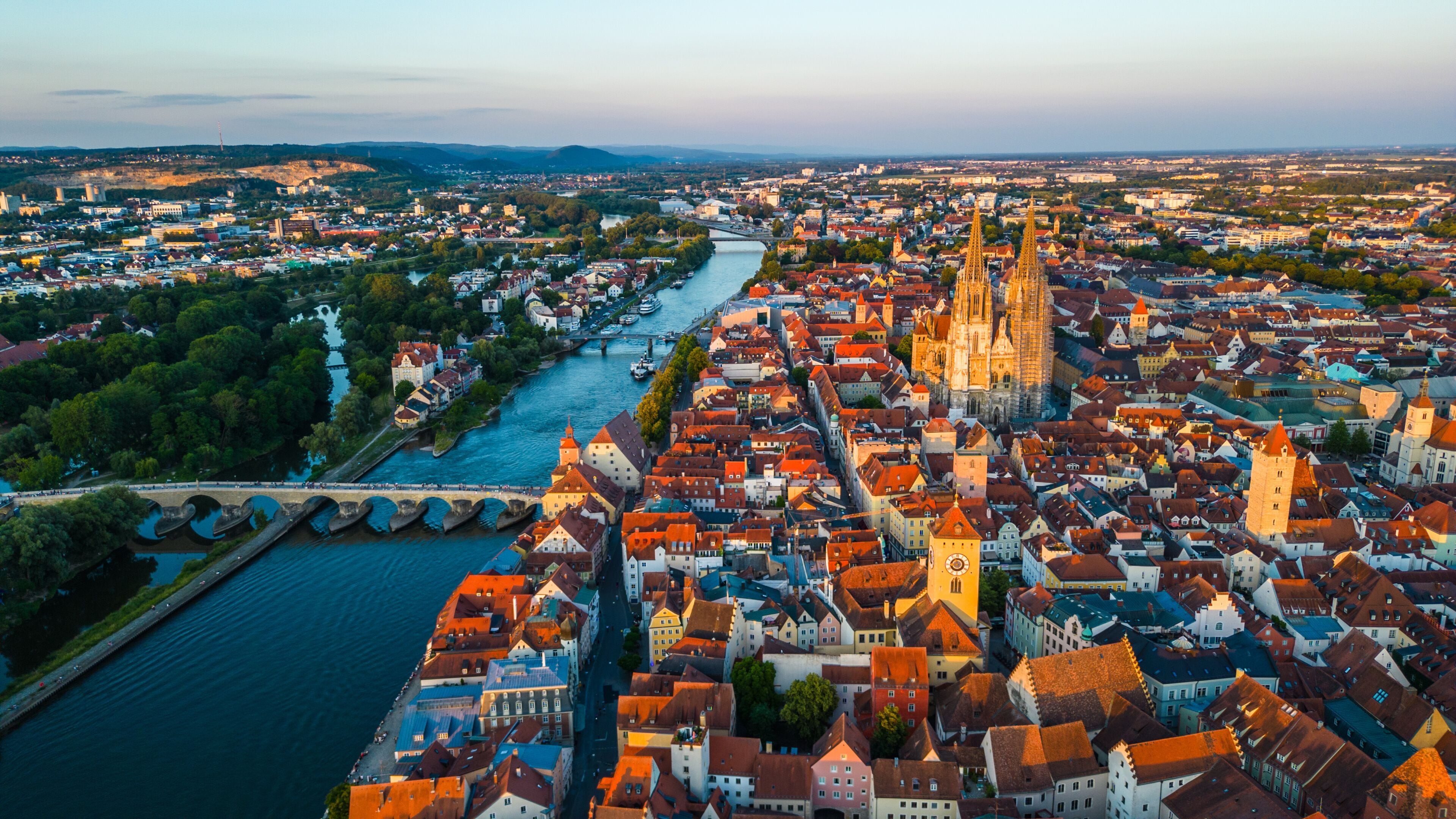 Regensburg at sunset captured from above, featuring the iconic Stone Bridge over the Danube River and the majestic twin towers of St. Peter's Cathedral glowing in warm evening light