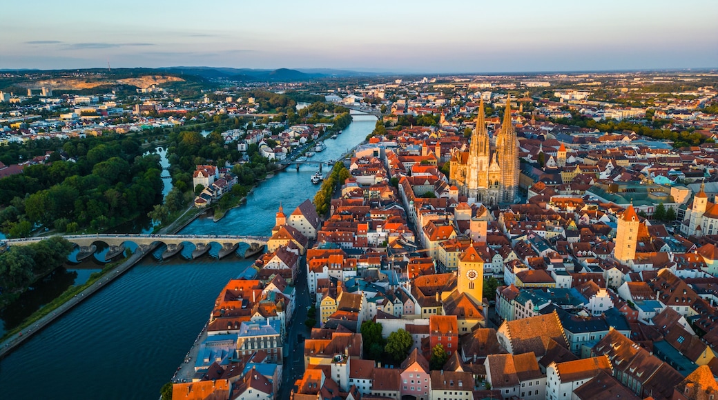 Regensburg at sunset captured from above, featuring the iconic Stone Bridge over the Danube River and the majestic twin towers of St. Peter's Cathedral glowing in warm evening light