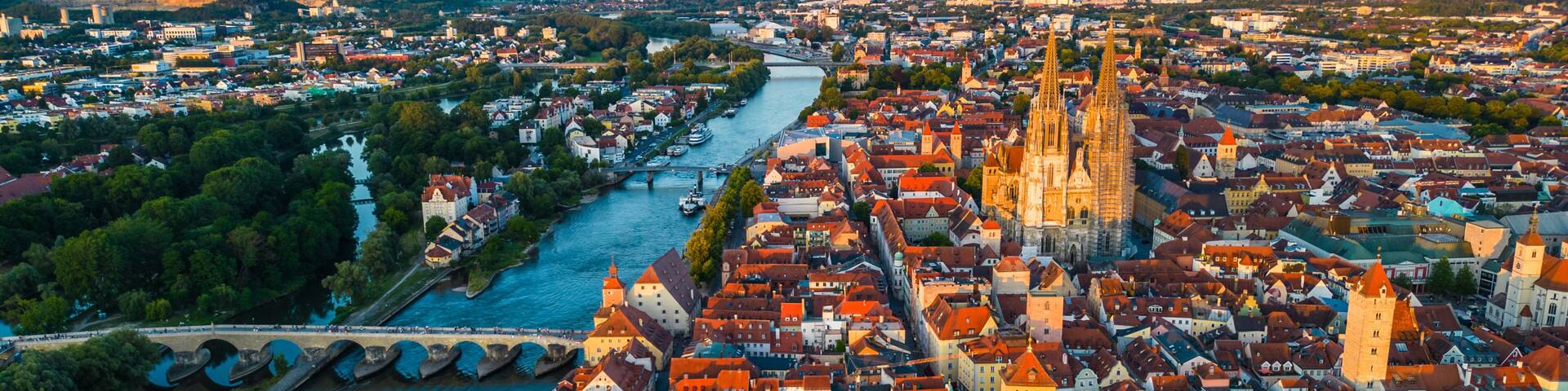 Regensburg at sunset captured from above, featuring the iconic Stone Bridge over the Danube River and the majestic twin towers of St. Peter's Cathedral glowing in warm evening light