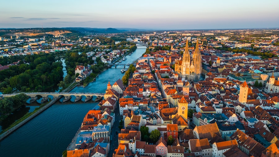 Regensburg at sunset captured from above, featuring the iconic Stone Bridge over the Danube River and the majestic twin towers of St. Peter's Cathedral glowing in warm evening light