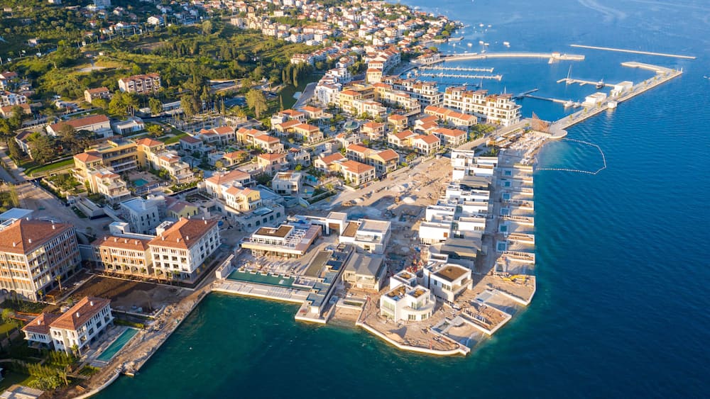 An aerial view of a coastal town with dense housing, a marina, and a clear blue sea.