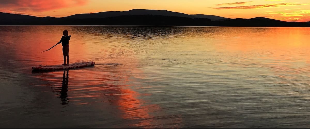 A person paddleboarding on calm water during sunset with mountains in the background.