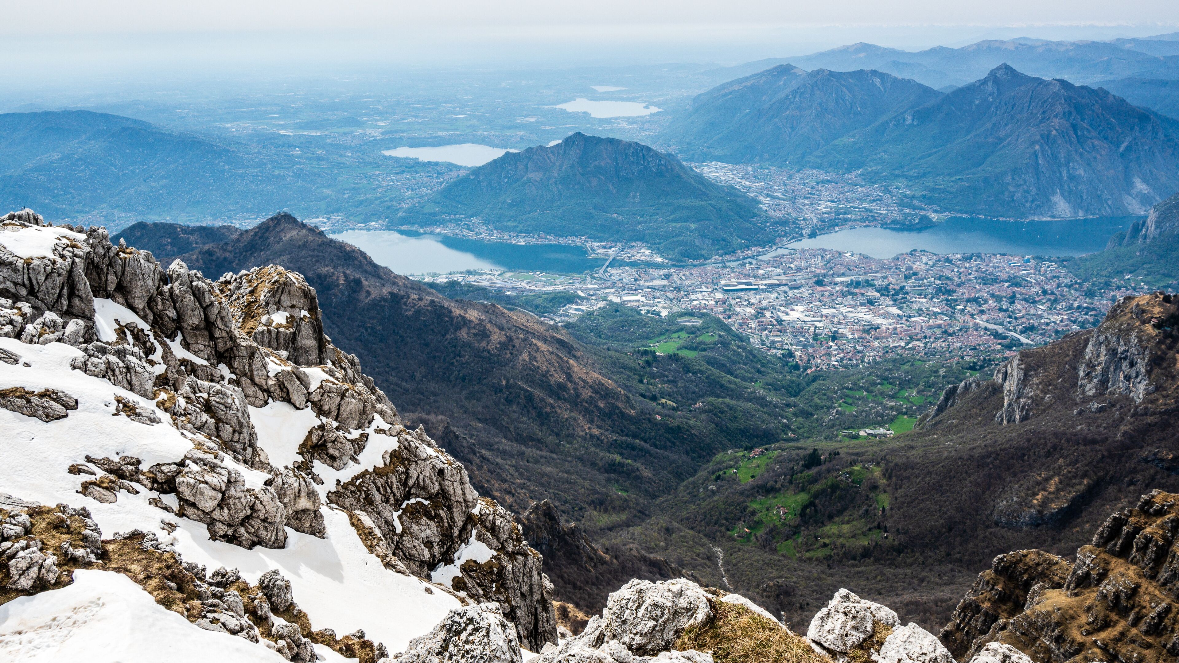 A mountainous landscape with rocky peaks, snow patches, and a distant cityscape with a lake.