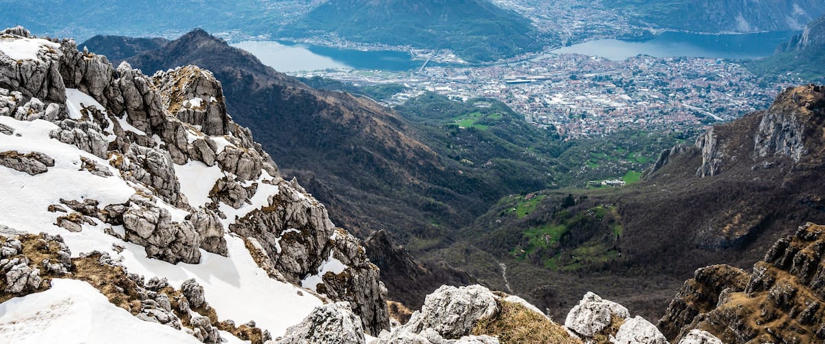 A mountainous landscape with rocky peaks, snow patches, and a distant cityscape with a lake.