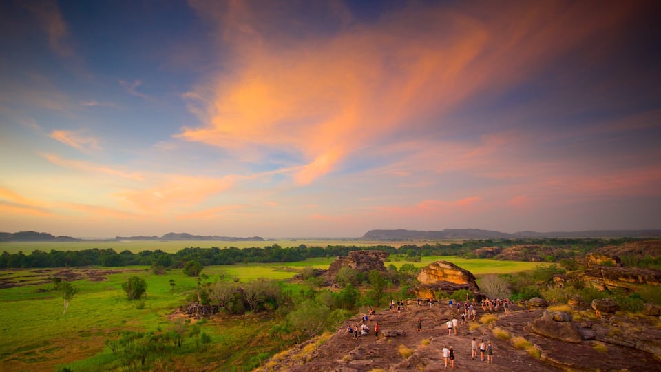 A landscape with a group of people on a rocky outcrop, surrounded by green fields and distant hills under a colorful sky.