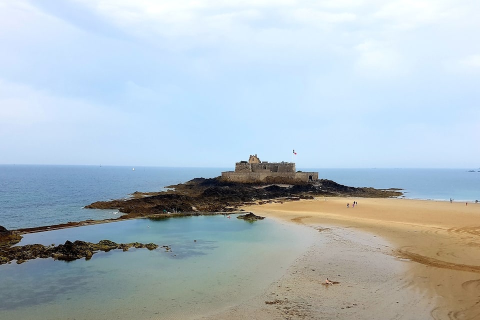 A coastal landscape with a historic fort on a rocky outcrop, a sandy beach, and calm sea.