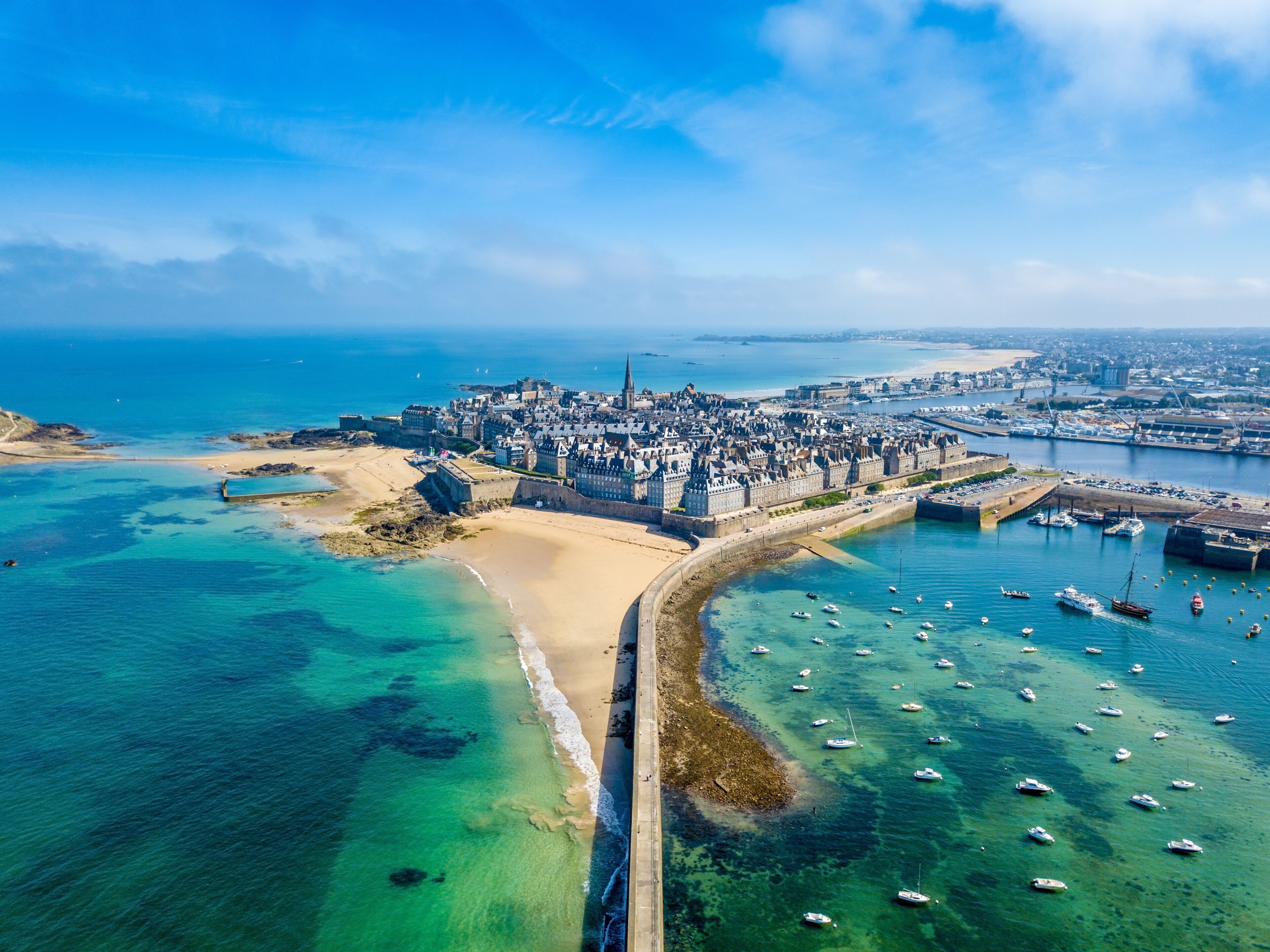 An aerial view of a coastal city with a prominent castle, a sandy beach, and a marina with numerous boats.