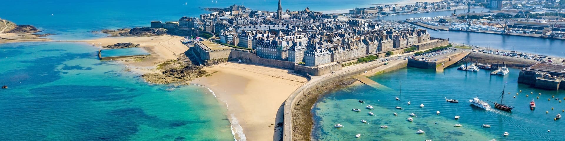 An aerial view of a coastal city with a prominent castle, a sandy beach, and a marina with numerous boats.
