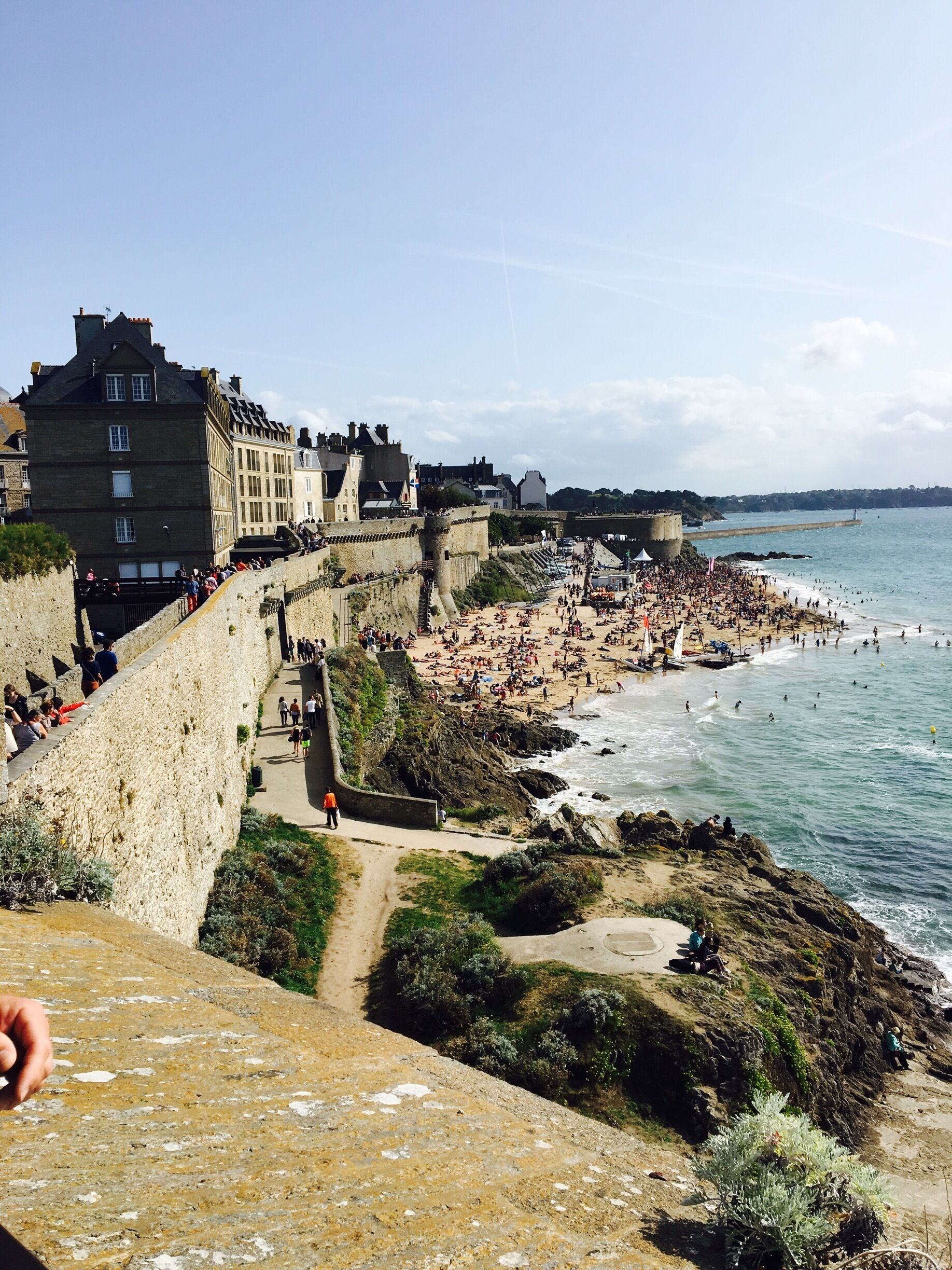 A coastal town with a historic stone wall, a crowded beach, and a clear blue sky.