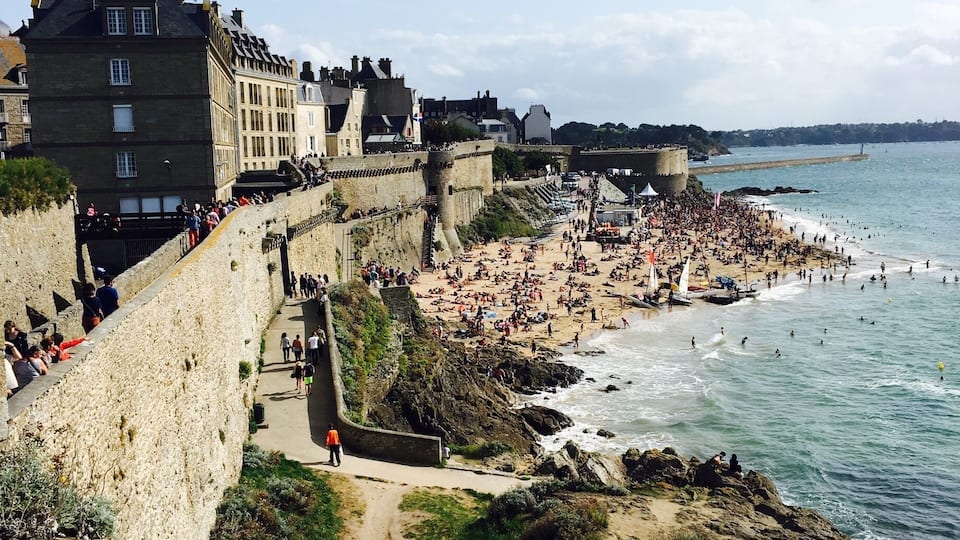 A coastal town with a historic stone wall, a crowded beach, and a clear blue sky.