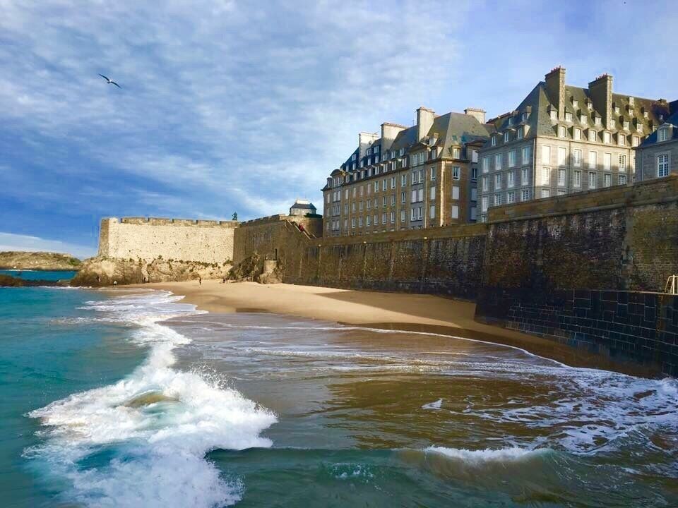 A beach with waves, a stone wall, and a large building with multiple windows.
