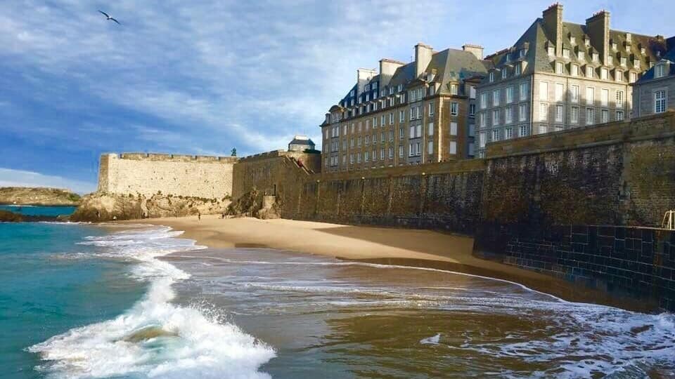 A beach with waves, a stone wall, and a large building with multiple windows.