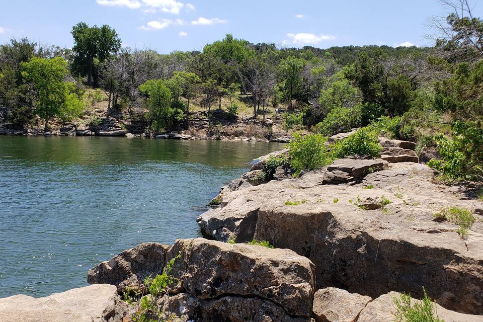 A rocky shoreline with a calm lake, surrounded by dense trees and a clear sky.