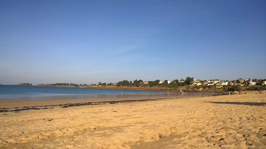 A sandy beach with a clear blue sky, calm sea, and houses in the background.