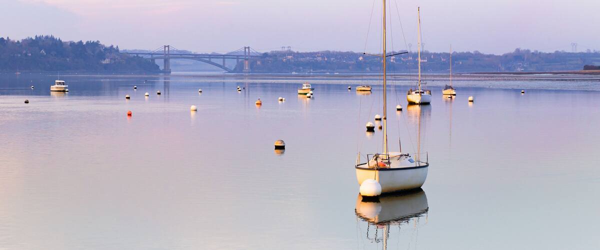 A calm harbor with several boats anchored near a bridge.