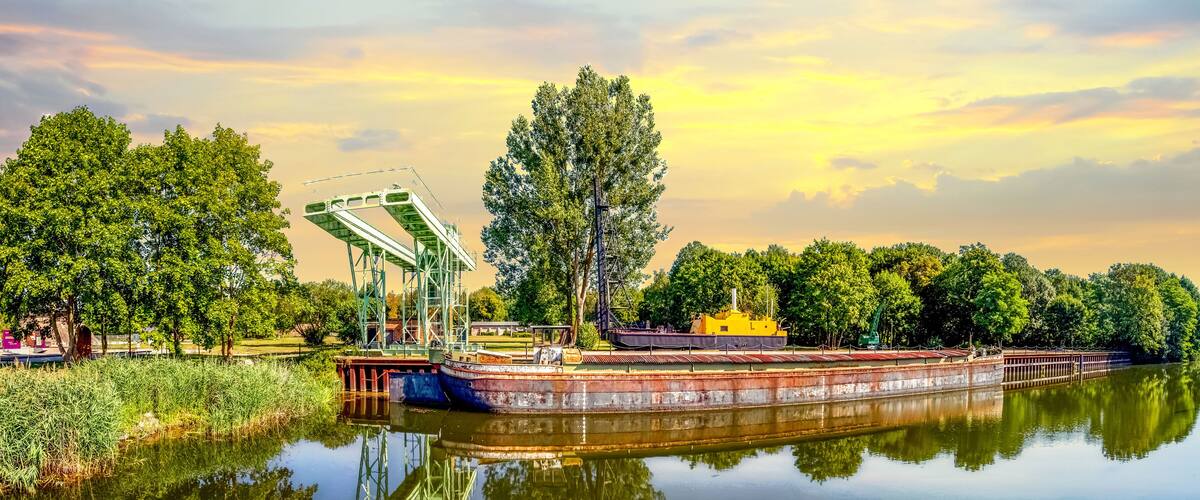 A calm river with a large barge docked on the right, surrounded by lush greenery and trees.