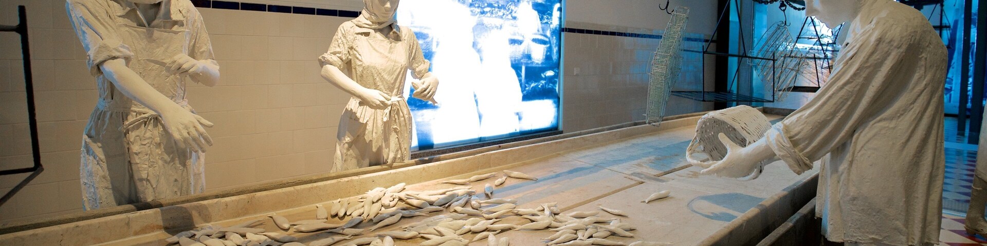 A museum exhibit with three white statues of figures in traditional attire, surrounded by a display of fish on a counter.