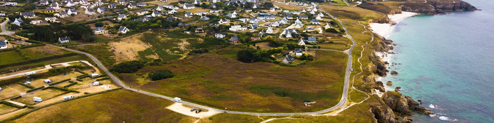 Vista aérea de un pueblo costero con casas, un camino serpenteante y acantilados rocosos.